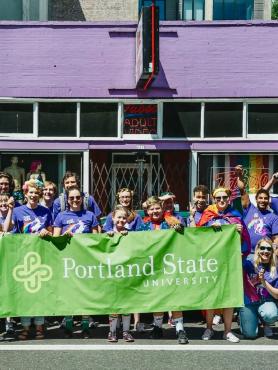 a large group of PSU affiliated people war matching purple shirts and hold a green PSU sign outside Stag waiting for the Pride Parade to begin