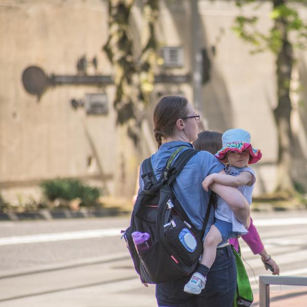 A mother with a backpack on walking outside while holding her young child.