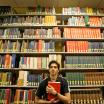 A student standing in front of tall library shelves holds a red bound book to their torso